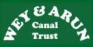 Wey and Arun Canal Trust Logo. Cream lettering "WEY & ARUN" in a semi circle above the words "Canal Trust" also in cream. All on a dark green background.