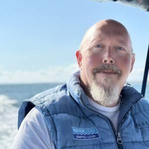 Ian Watkins, Waterlife. A bald man with a goatee beard wearing a blue gilet on board his boat with the sea in the background. 