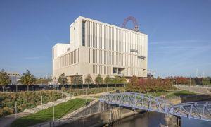 UCL East, Marshgate. A modern, cream coloured concrete building with an iron bridge over the Waterworks River in the foreground. Above the roof can be seen the red framework of the top of the ArcelorMittal Orbit.