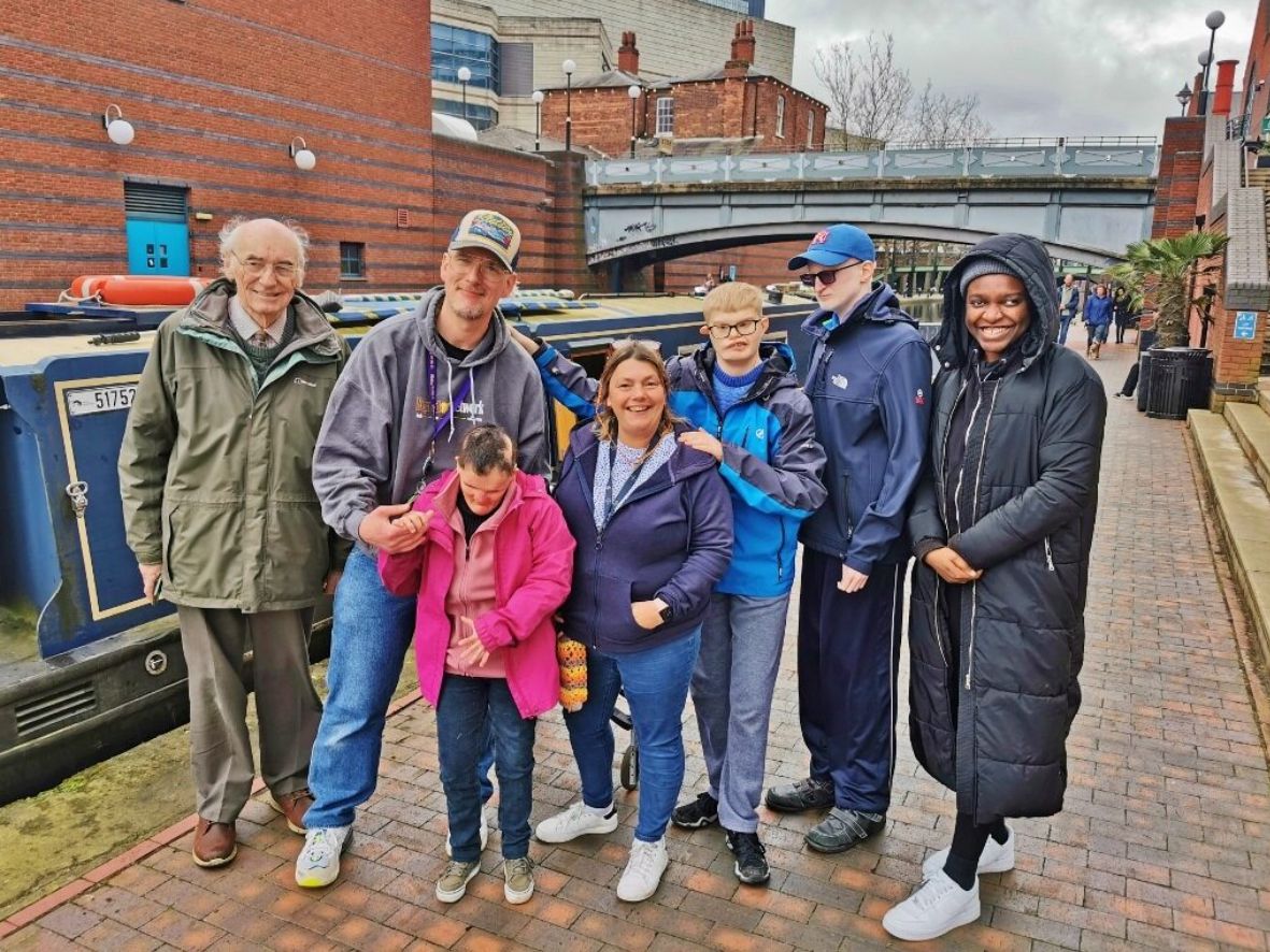 A group of seven people are standing on a brick towpath next to a canal boat. The group consists of three adults with disabilities, two adults who appear to be carers, and two young men. They are all smiling and looking at the camera. The photo has been taken on the canal in central Birmingham.